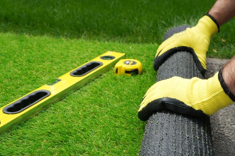 Synthetic-Turf-Installation-mans-hands-spread-artificial-turf-roll Close-up of Lawnmax worker laying artificial grass turf with gloves, spirit level and tape measure